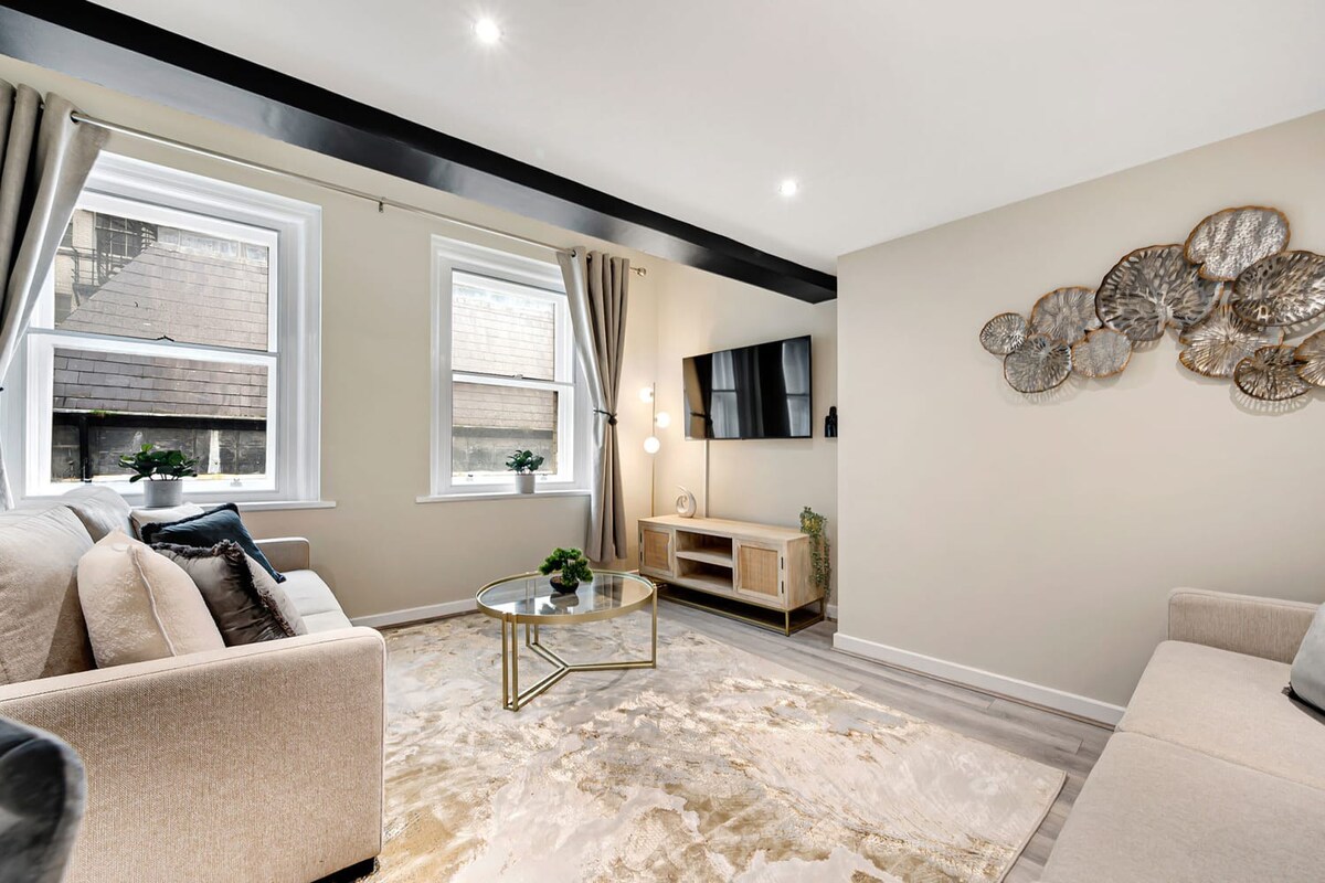 A bright living room with a light beige sofa, a wall-mounted TV above a wooden media console, and a gold-framed coffee table on a patterned rug, with natural light entering through two large windows.