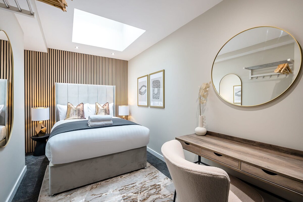 A modern bedroom with a large bed featuring an upholstered headboard against a slatted wood accent wall, a wooden desk with a light chair, and a large round mirror, all beneath a rectangular skylight.