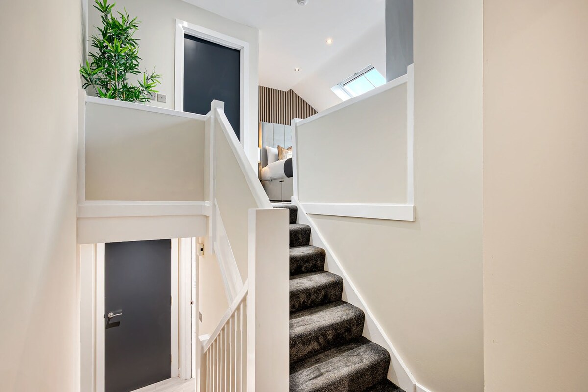 A modern interior view of a carpeted staircase with white railings and light beige walls, leading up to a bedroom with a slatted wood accent wall and a skylight, and down to a dark door.