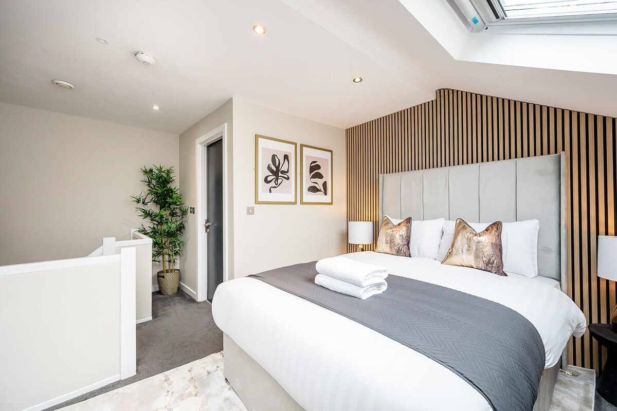 A bright bedroom featuring a large bed with a grey upholstered headboard against a vertical slatted wood accent wall, two framed art pieces, and a skylight, with light grey carpet.