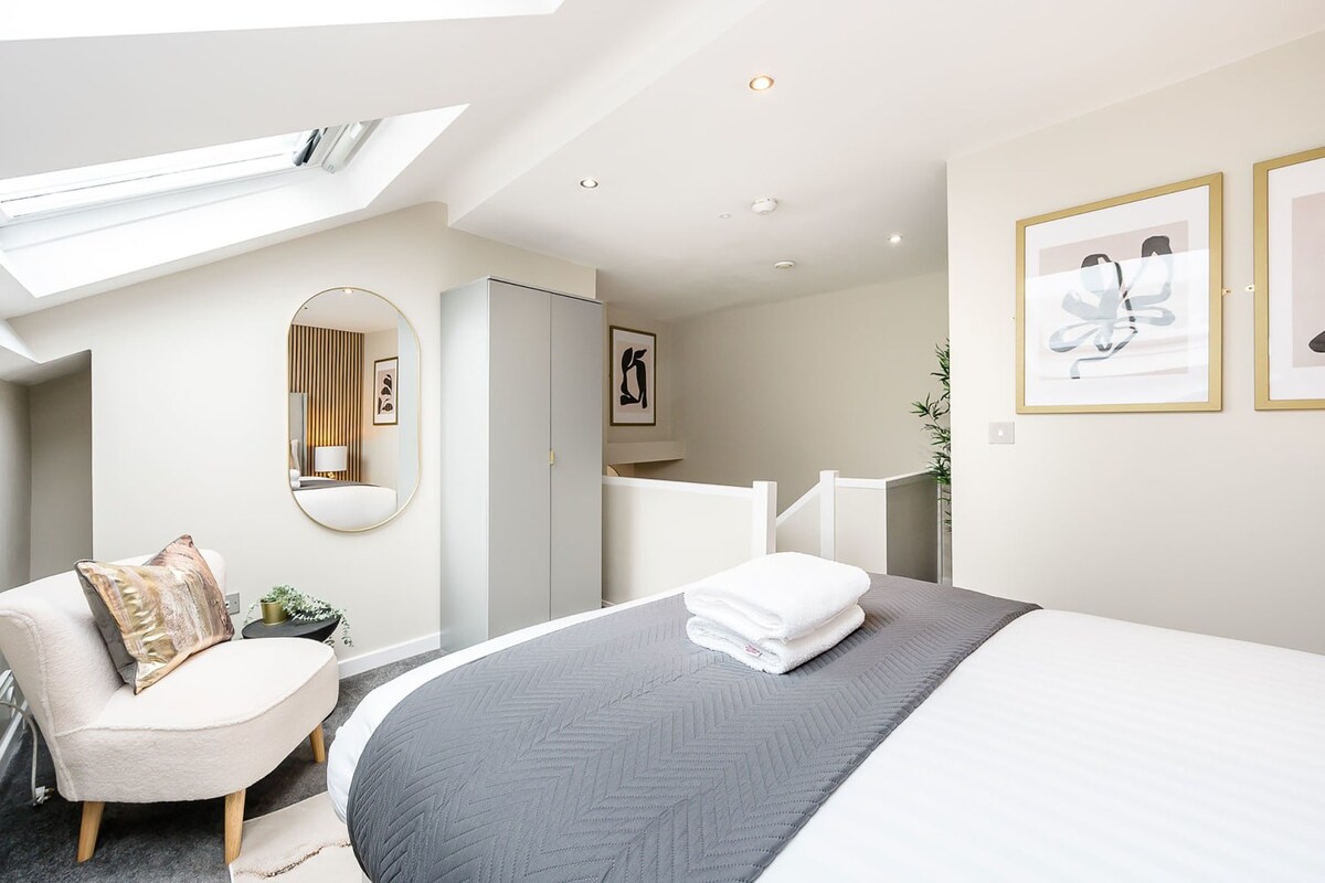 A cozy bedroom with a large bed, a cream accent chair, a full-length oval mirror, and a grey wardrobe, all under a sloped ceiling with a large skylight.