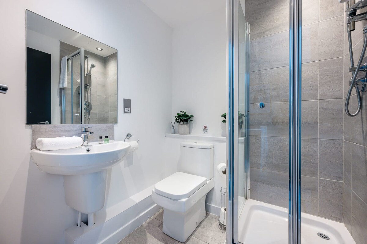 A modern bathroom with a white pedestal sink, toilet, and a glass-enclosed shower with grey tiled walls, featuring a large mirror above the sink.