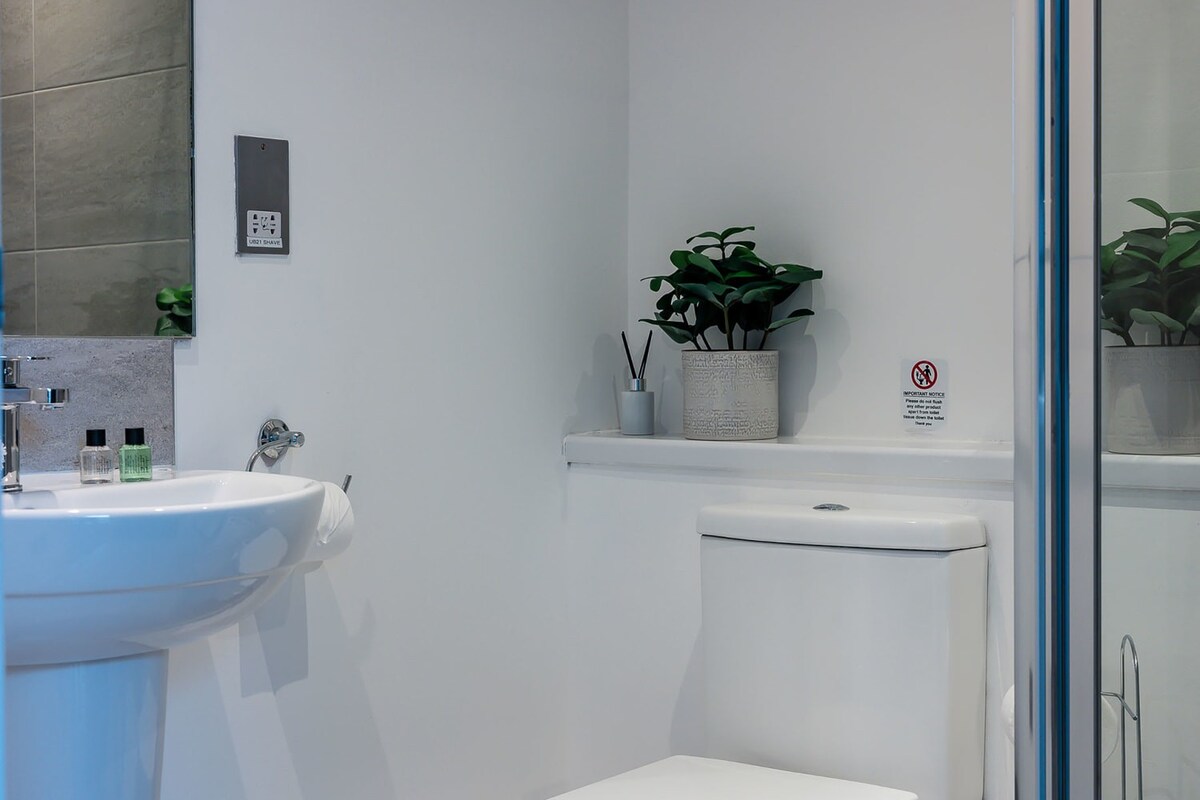 A close-up view of the modern bathroom's white toilet and sink area, featuring a small potted plant and diffuser on a shelf above the toilet, and a mirror reflecting the shower.