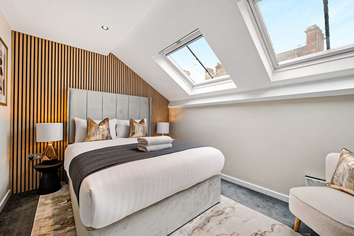 A bright attic bedroom with a large bed, a grey upholstered headboard against a slatted wood accent wall, and two large skylight windows offering views of the outside.