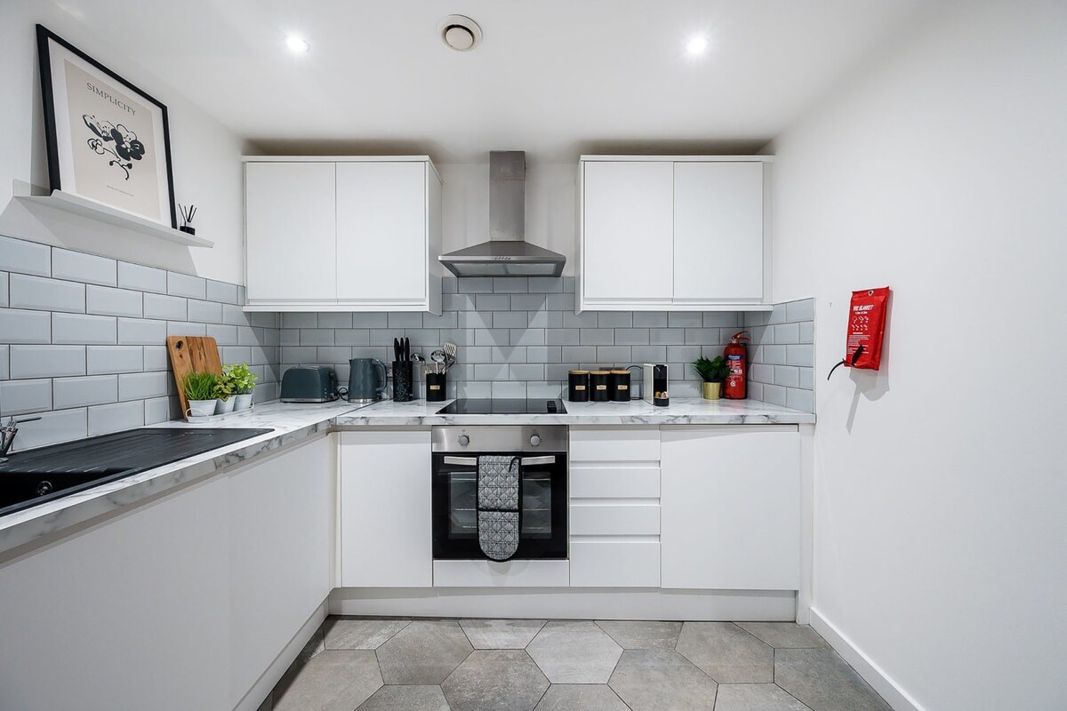 A modern white kitchen with L-shaped cabinetry, light grey subway tile backsplash, marble-effect countertops, and stainless steel appliances, including an oven and range hood.
