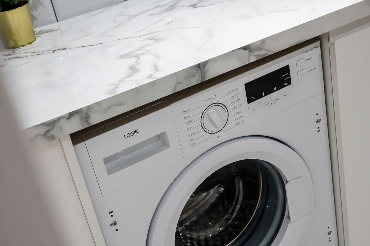 A close-up of a white Logik washing machine integrated under a marble-effect countertop in a modern kitchen.
