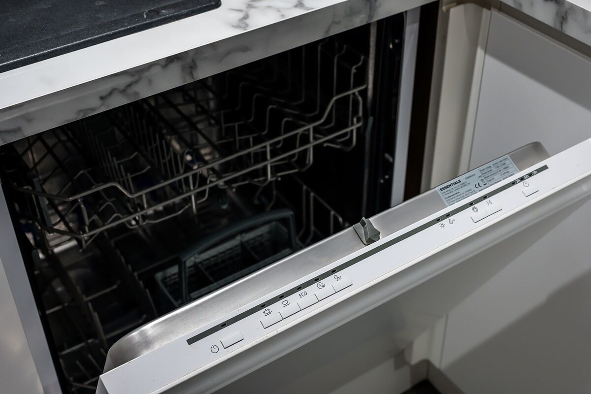 A close-up of an integrated Essentials dishwasher with its door ajar, revealing the racks inside, set under a marble-effect countertop in a white kitchen.