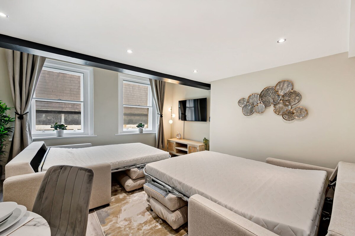 A modern living space featuring two pulled-out sofa beds with white mattresses, light-colored walls, and two large windows with grey curtains. A flat-screen TV is mounted on the wall, complemented by a decorative gold metal wall art piece and recessed lighting.