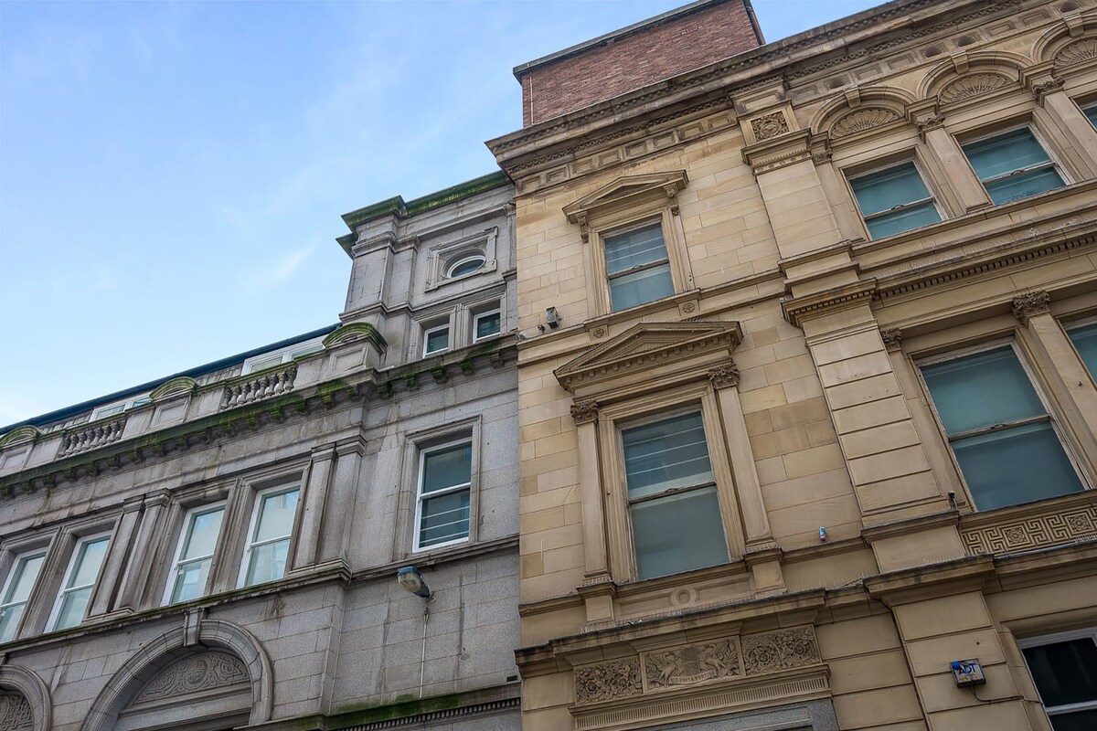 A close-up, upward-angled view of the upper sections of two adjacent historic buildings, showcasing their detailed stone facades, multiple windows, and contrasting architectural styles under a clear sky. The building on the right features more elaborate carvings and a lighter stone.