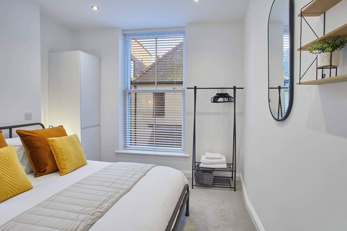 A bright bedroom features a metal-frame bed with white bedding and mustard yellow and rust-colored pillows, a large window with white blinds, and a minimalist black clothes rack with folded towels. A tall white wardrobe stands in the corner.