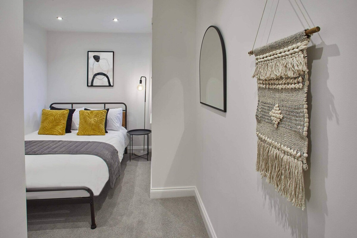 A cozy bedroom with a metal-frame bed dressed in white and grey bedding, accented by mustard yellow pillows. An arched black mirror and a woven wall hanging adorn the light grey wall, with a modern floor lamp visible in the background.