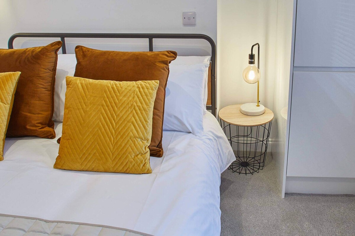 A close-up of a bed with white bedding and vibrant mustard yellow and rust-colored pillows, featuring a modern bedside table with a light wood top and black wire base, illuminated by a stylish lamp with an exposed bulb.