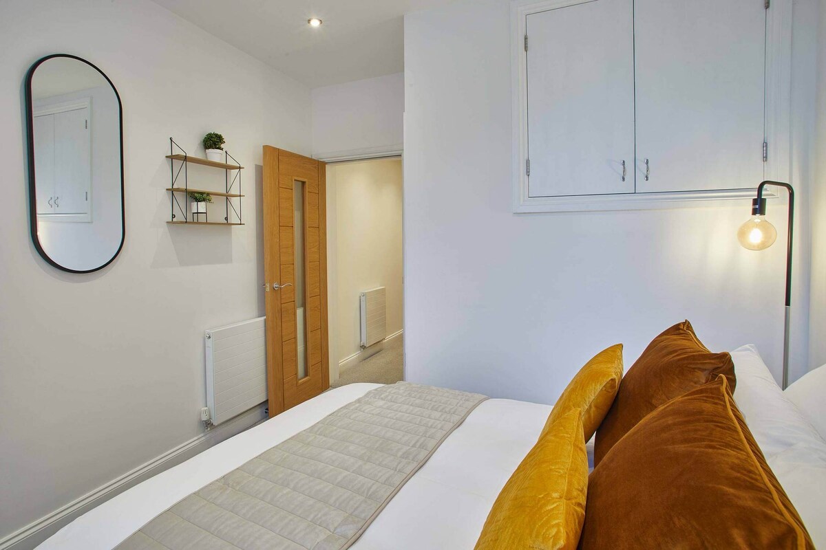 A bedroom view showcasing a metal-frame bed with white bedding and warm-toned pillows, an oval black-framed mirror, and a small wall-mounted shelf with greenery. A wooden door and a modern bedside table with a lamp are also visible.