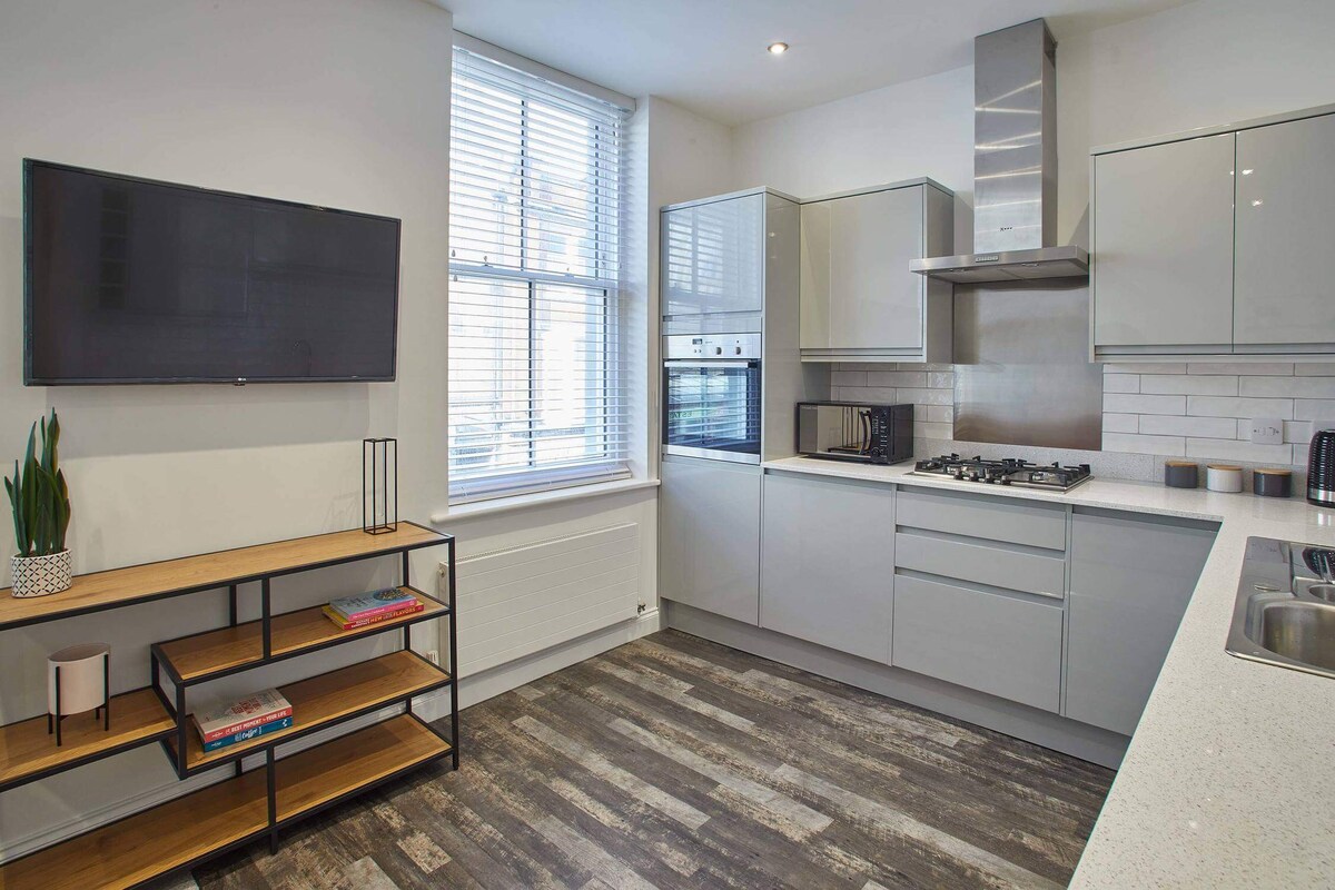 Modern kitchen area with a wall-mounted TV above a wooden and metal shelving unit, next to a large window, showcasing light grey cabinetry and stainless steel appliances.