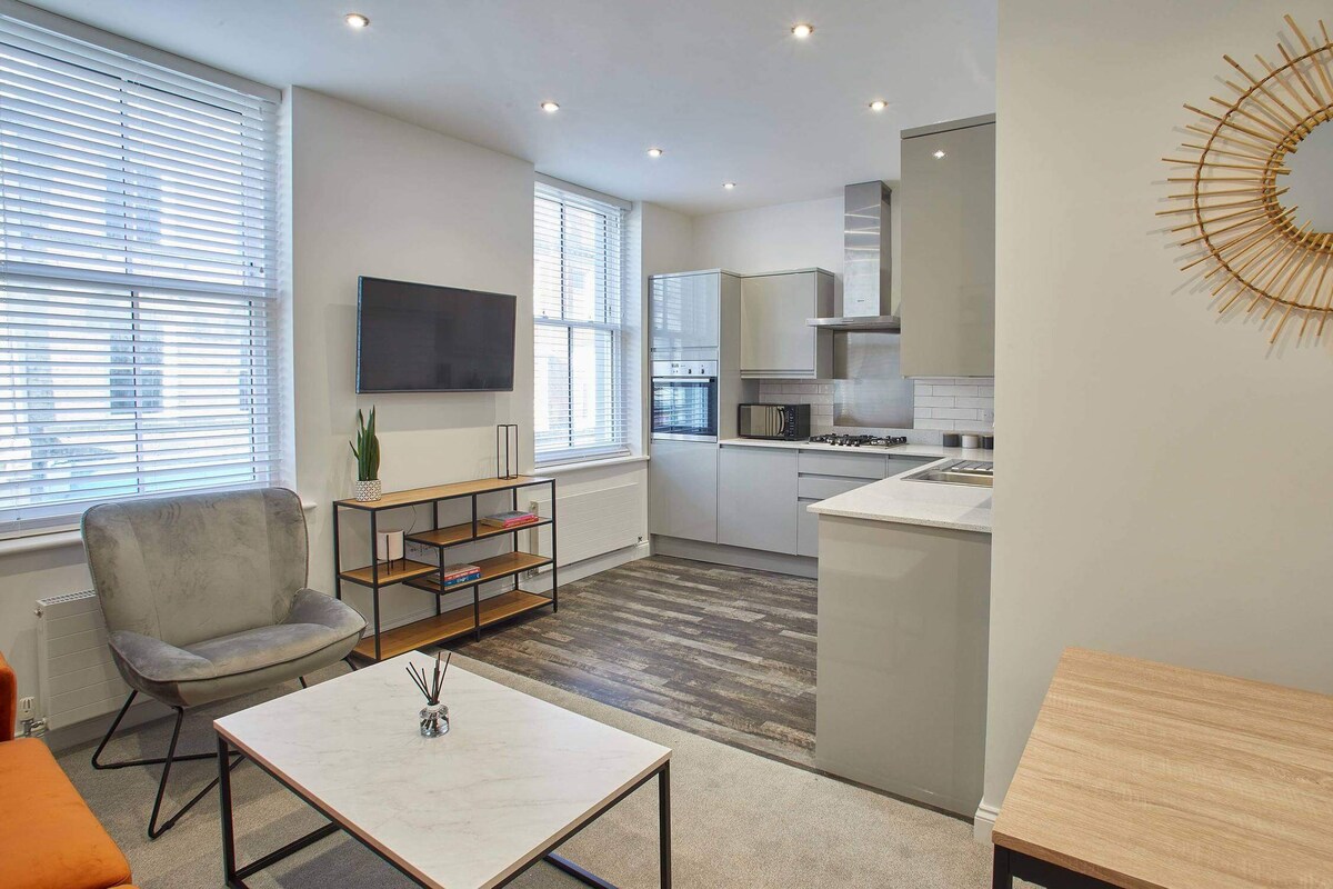 View of the living room with a wall-mounted TV and a grey armchair, transitioning into the modern kitchen equipped with light grey cabinets and stainless steel appliances.