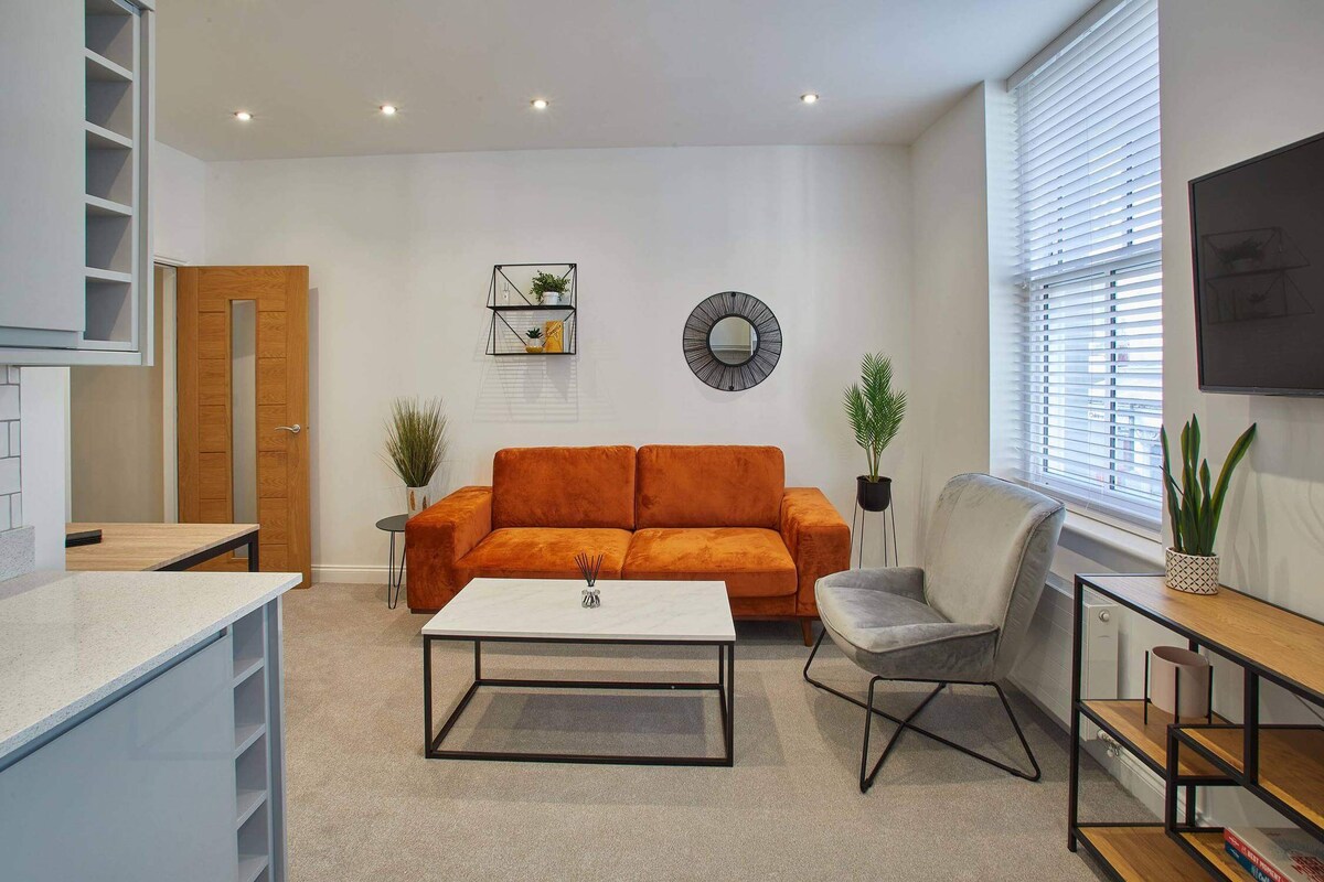 Open-concept living space with an orange velvet sofa, a grey accent armchair, and a white marble-top coffee table, flowing into the kitchen and dining area.