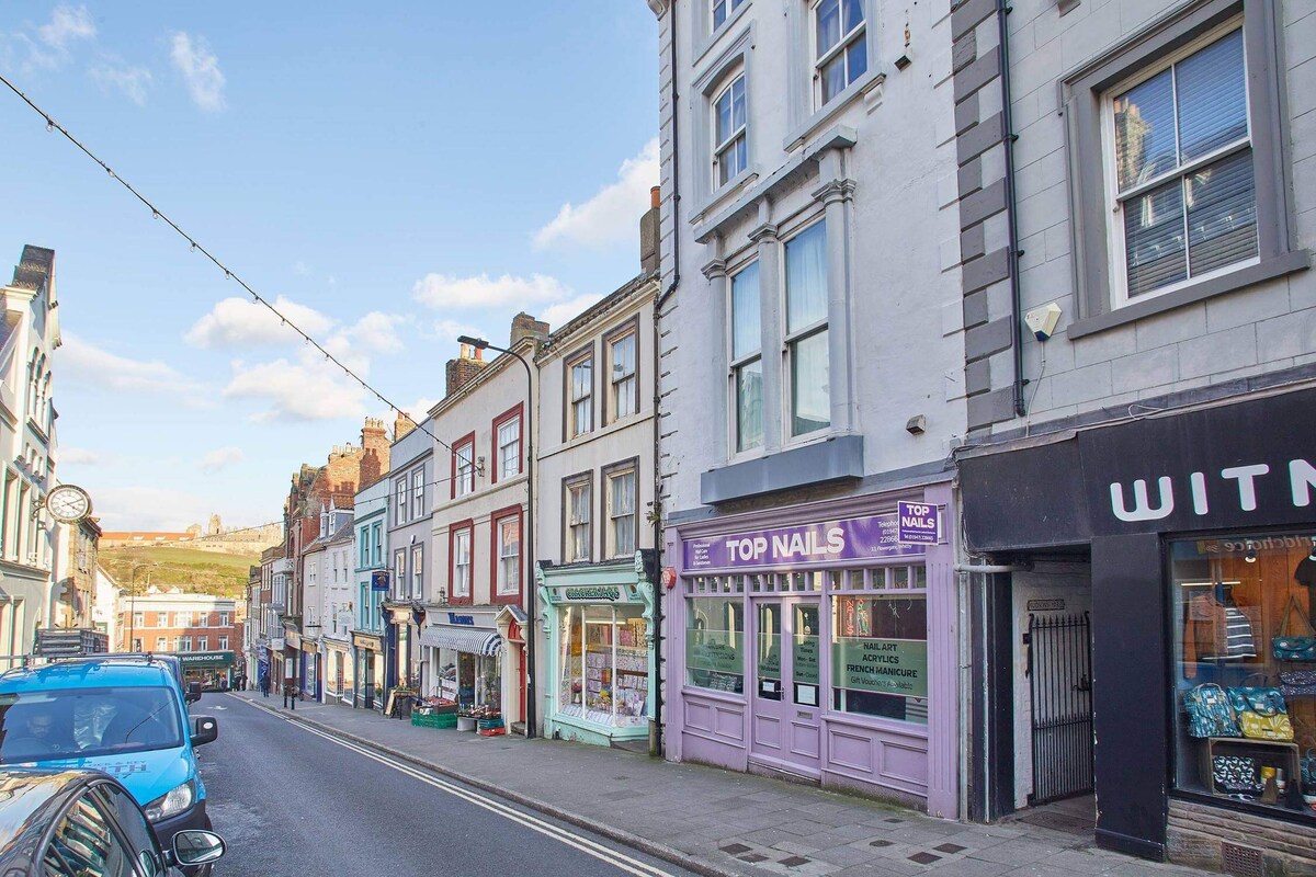 A vibrant street scene with a row of historic buildings housing various shops, including a "Top Nails" salon with a purple facade, under a bright blue sky with scattered clouds. Parked cars line the street.