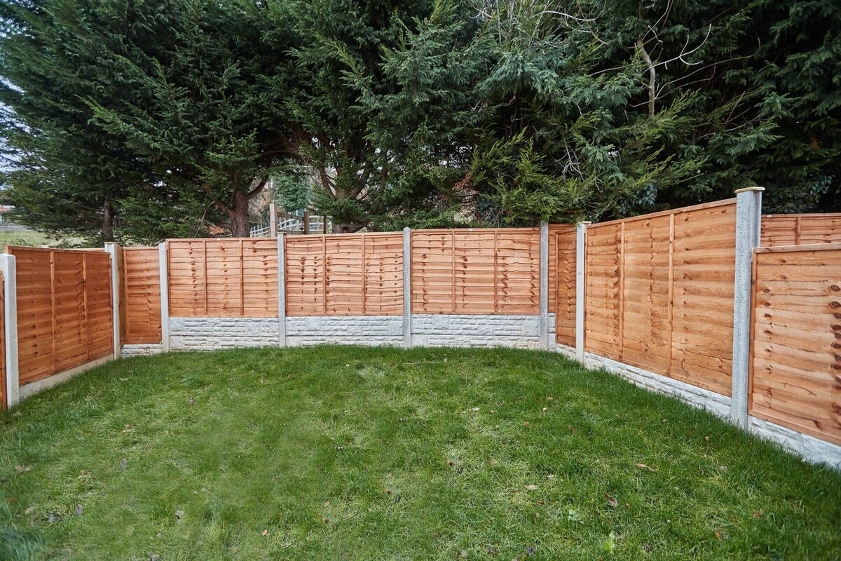A well-maintained, rectangular grassy yard enclosed by new wooden fence panels with concrete bases, backed by a dense line of tall evergreen trees providing privacy.