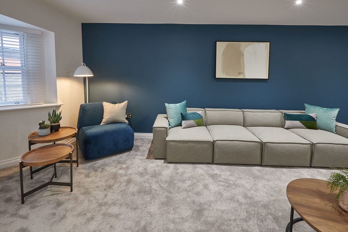 A cozy corner of the living room featuring a light grey modular sectional sofa and a blue accent chair with a floor lamp. Two small wooden side tables hold potted plants, and a window with white blinds is visible, all set against a deep blue accent wall with abstract art and grey carpet.