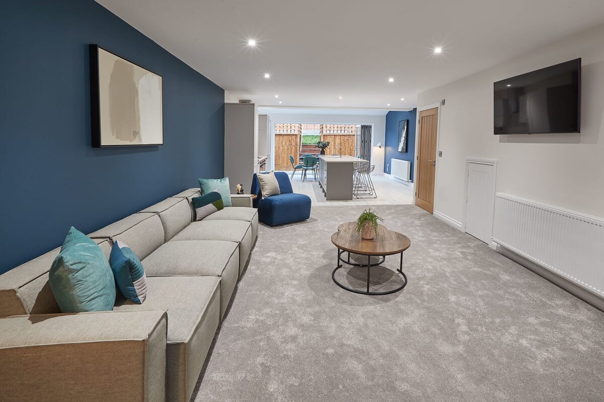 A wide view of the open-plan living room, featuring a large light grey sectional sofa, two round wooden coffee tables, and a wall-mounted TV. A deep blue accent wall provides a focal point, and the room transitions from grey carpet to light wood-effect flooring towards the kitchen and dining area.