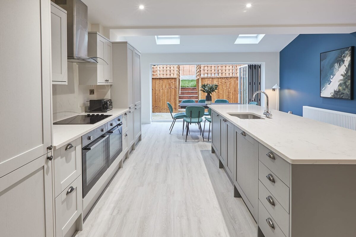 A modern kitchen with light grey shaker-style cabinets, white quartz countertops, and integrated stainless steel appliances including an oven, hob, and range hood. A toaster and kettle are visible on the counter, and the room features light wood-effect flooring and skylights, leading to the dining area.