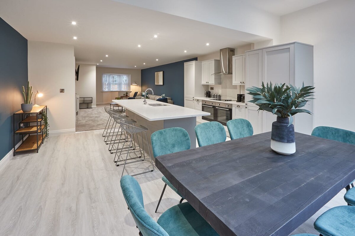 An expansive open-concept space showcasing the kitchen island with bar stools, a dark wooden dining table with teal chairs, and the living room with a large grey sofa in the background. A deep blue accent wall adds color, and the flooring transitions from light wood-effect to grey carpet.