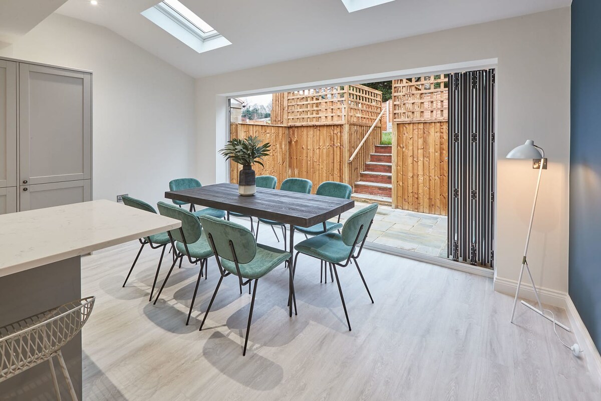 A bright dining area featuring a dark wooden table with eight teal velvet chairs, illuminated by natural light from skylights and bi-fold doors that open to an outdoor patio. A modern white floor lamp stands in the corner, and a plant centerpiece adorns the table, all on light wood-effect flooring.