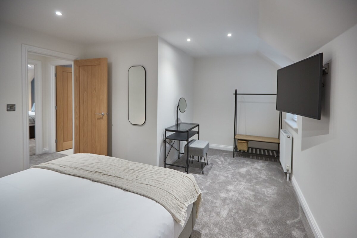 Bright bedroom with light grey carpet, white walls, and a bed with white linens. Features a wall-mounted TV, a black metal dressing table with a round mirror, and an open clothes rack.