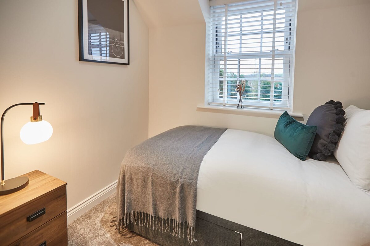 Another view of the single bedroom, showcasing a dark wood bedside table with a modern lamp. The bed is dressed in white linens with a grey throw and accent pillows, next to a window with white blinds.