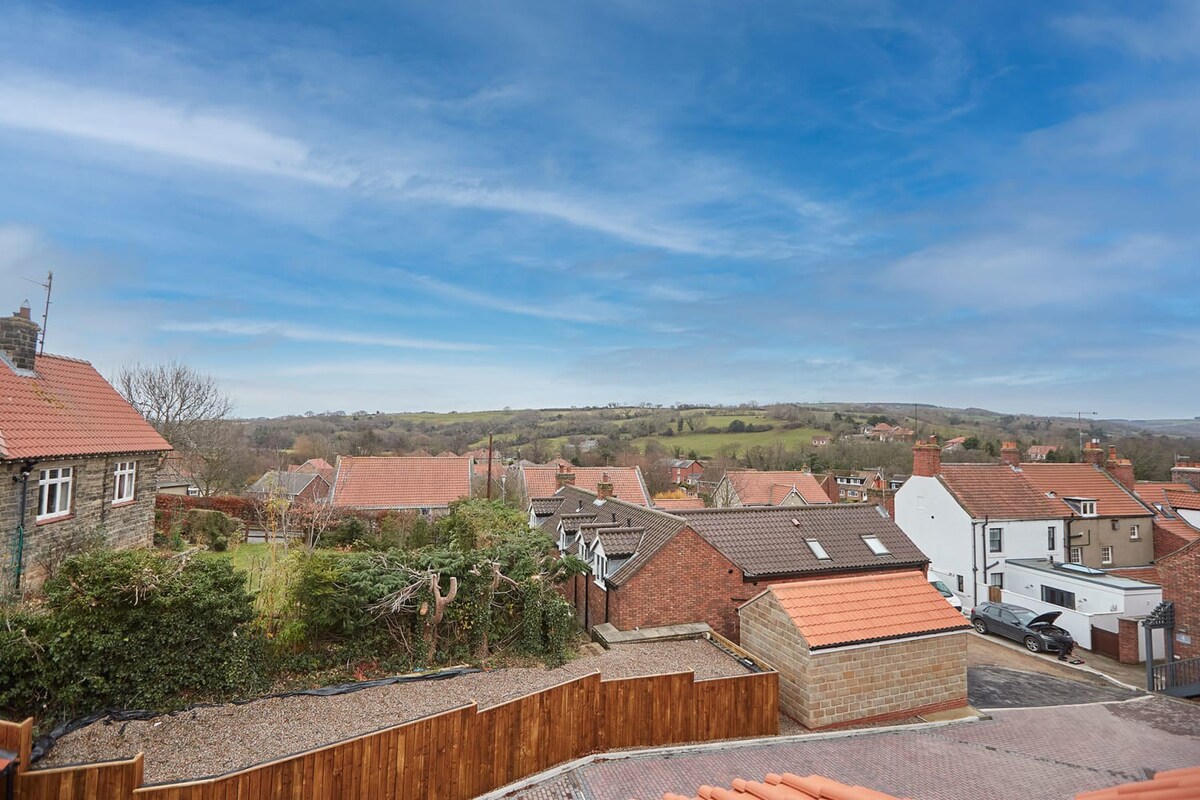 An elevated view of a charming village with terracotta-roofed houses nestled among rolling green hills under a bright blue sky with wispy clouds, showcasing the rural landscape.