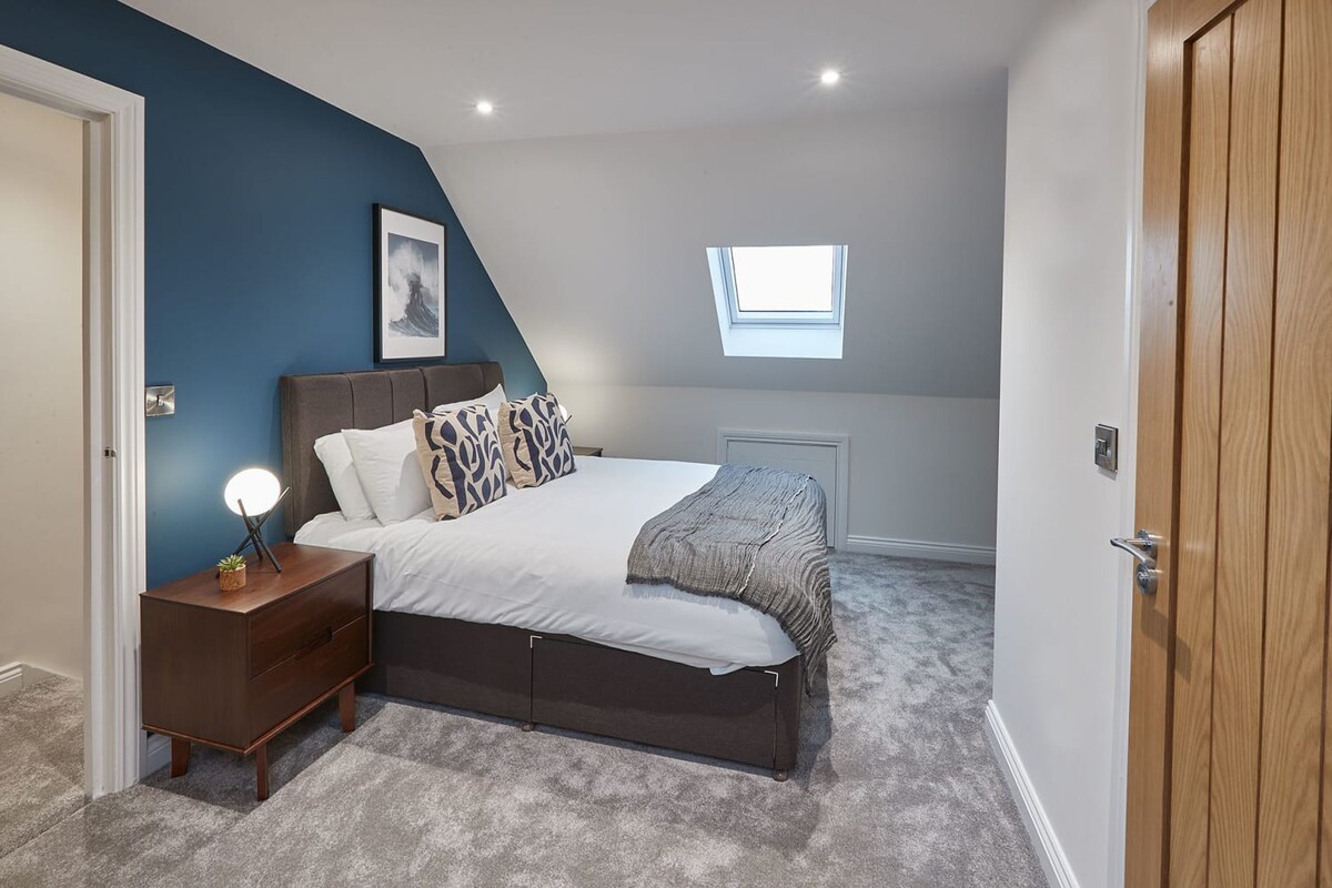 Wide shot of the bedroom with a double bed, a dark blue accent wall, and a sloped ceiling with a skylight. A wooden door is open, revealing a glimpse into the hallway, and a bedside table with a lamp is visible.