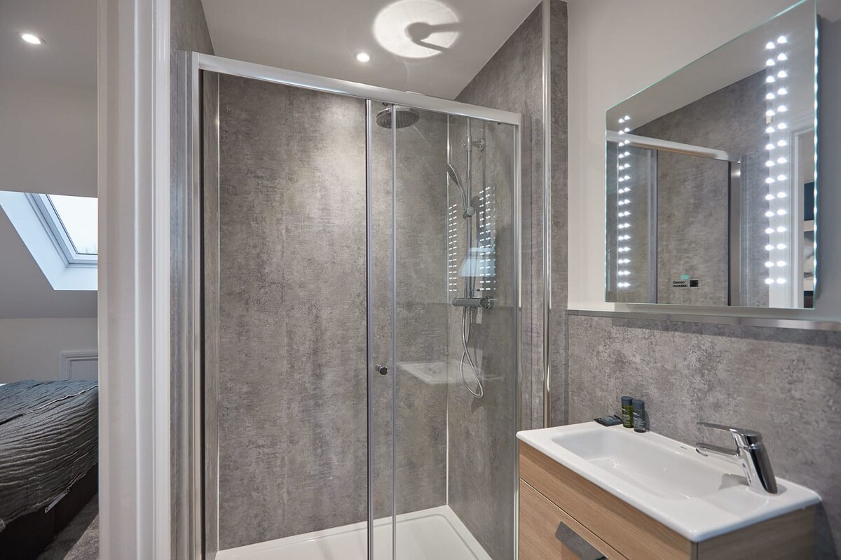 Compact ensuite bathroom with grey textured wall panels, featuring a glass shower enclosure, a light wood vanity with a white sink, and an illuminated mirror. A glimpse of a bedroom with a skylight is visible through the doorway.