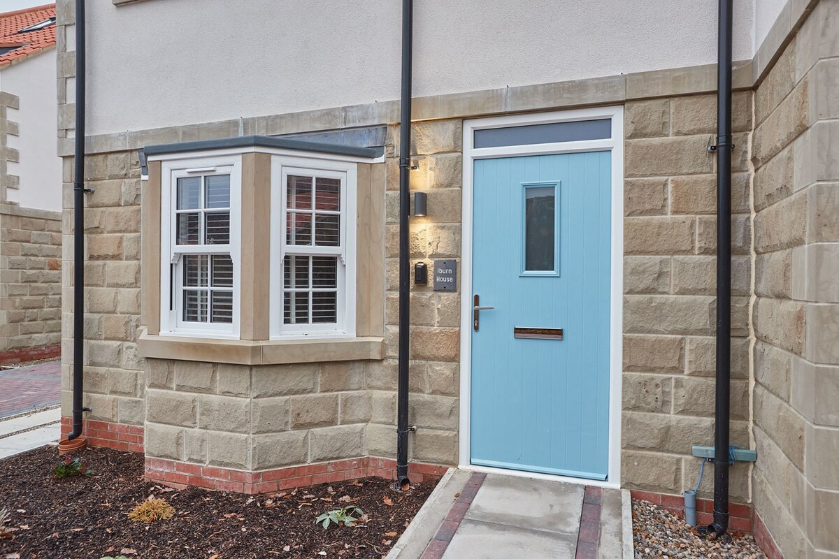 The inviting front entrance of a modern stone and stucco house, featuring a light blue paneled door with a small window, a white bay window, and a small nameplate reading 'Burn House'.