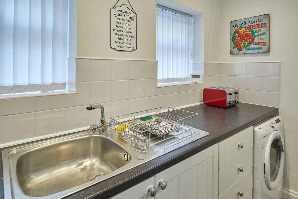Close-up of the kitchen sink area with a stainless steel sink, dish rack, and a washing machine integrated under the dark gray countertop. White shaker-style cabinets and vertical blinds on the windows complete the look.