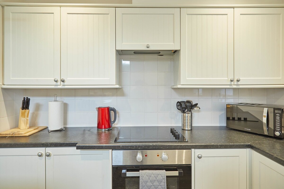 Close-up view of the kitchen showing the electric hob, built-in oven, and dark gray countertop. White shaker-style cabinets are above and below, with a red kettle and knife block on the counter.