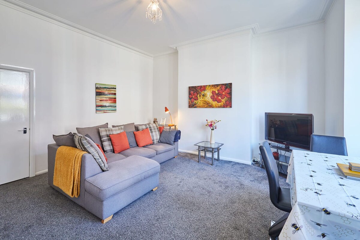 View of the living room showing the gray L-shaped sectional sofa with orange and plaid pillows, a small glass coffee table, and a dining table with a patterned tablecloth. A door is visible on the left, and artwork adorns the white walls.