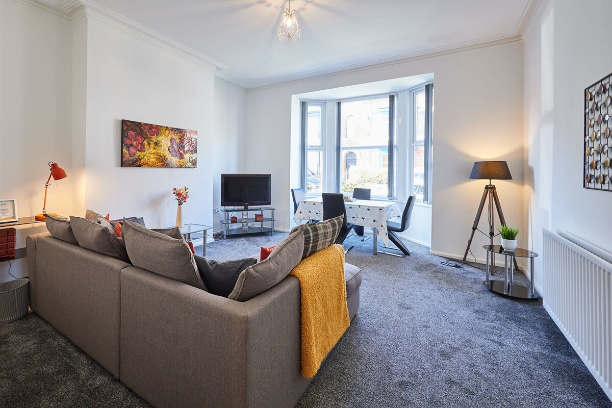 Spacious living room with a gray L-shaped sofa, a TV on a stand, and a dining area with a table and chairs in a bay window. The room features light gray carpet, white walls, and a mix of modern and abstract artwork.