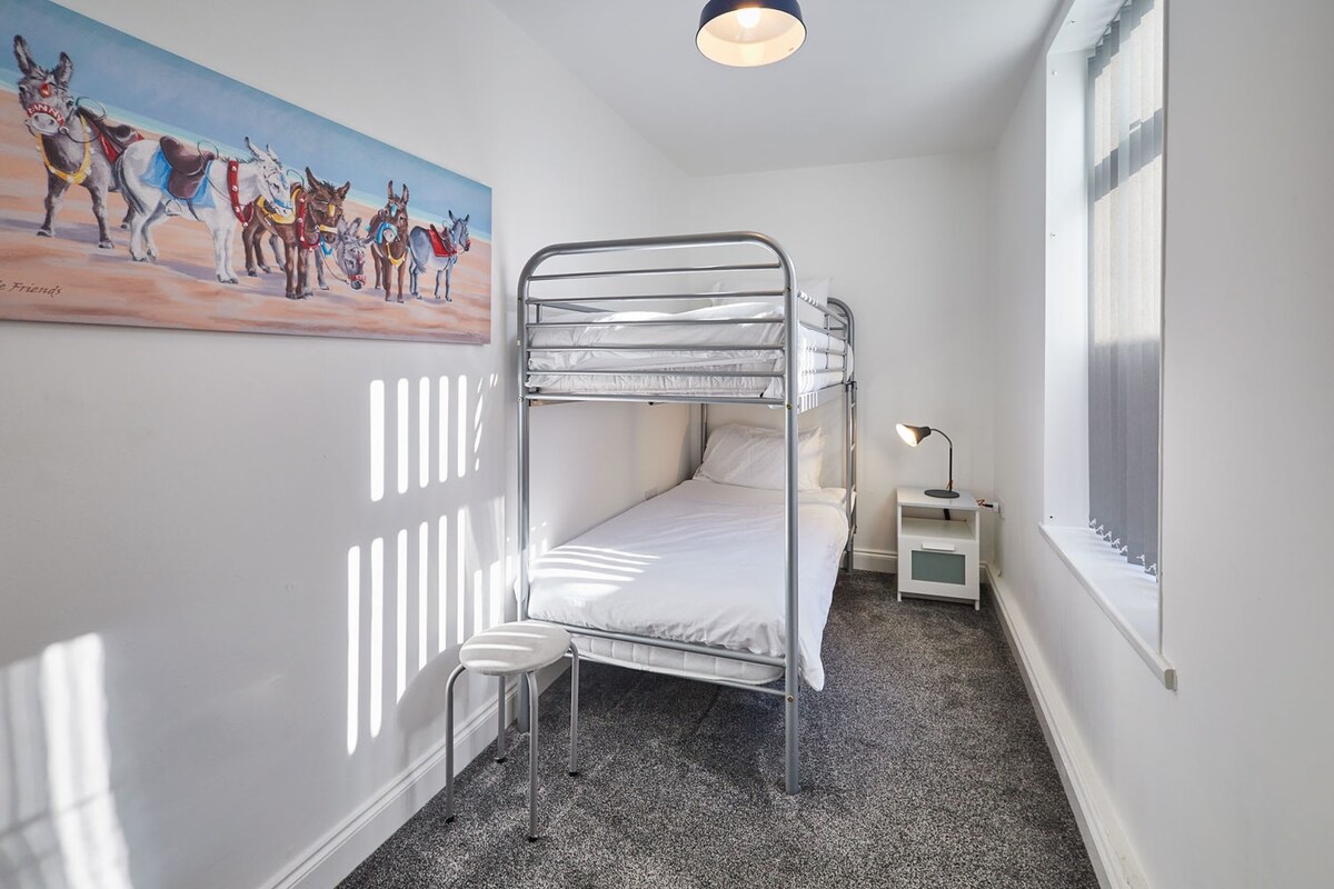 A narrow bedroom with white walls, dark grey carpet, and a silver metal bunk bed with crisp white bedding. A colorful donkey painting hangs on the wall, complemented by a small white nightstand with a lamp next to the bed.