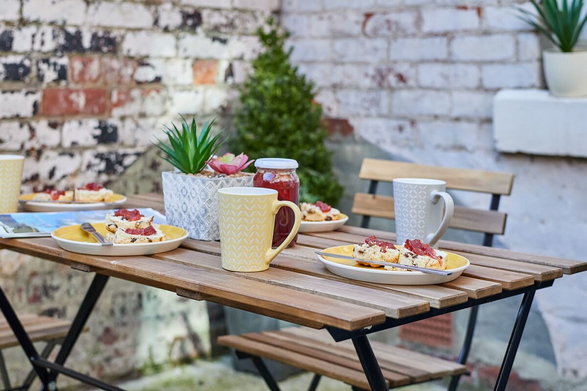 A close-up view of an outdoor wooden folding table laden with plates of scones topped with jam, mugs, and a jar of jam. Potted succulents and a textured brick wall are visible in the background, creating a cozy outdoor dining ambiance.