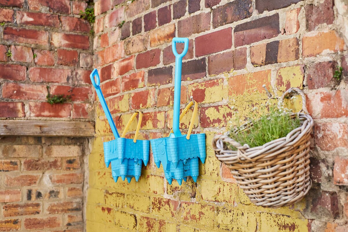 A rustic brick wall, partially painted yellow, serves as a backdrop for two decorative blue castle-shaped sand shovels. A woven basket filled with greenery is also mounted on the wall, adding a charming touch to the outdoor space.