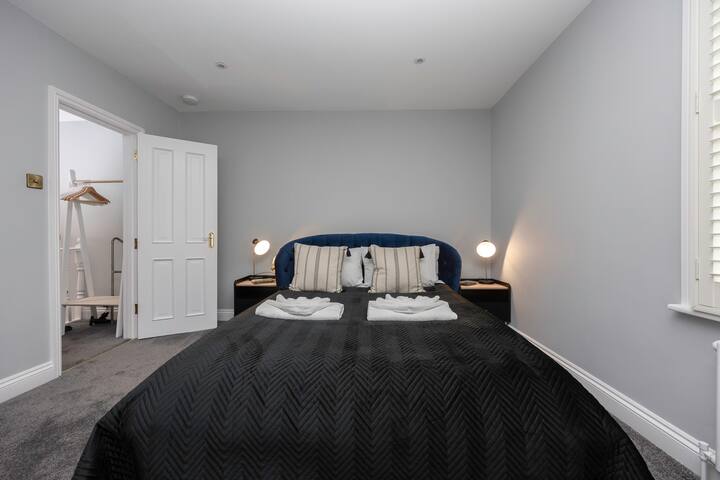 A modern bedroom with light gray walls, a dark blue upholstered headboard, a black quilted bedspread, and two white nightstands with small white lamps. A white door leads to a walk-in closet or dressing area.