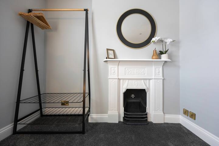 A corner of a bedroom featuring a decorative white fireplace with a black grate, topped by a round mirror with a black and gold frame, and an open metal and wood wardrobe on dark grey carpet.