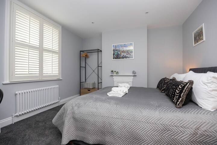 A bedroom with light gray walls and gray carpet, featuring a bed with a gray chevron-patterned bedspread and decorative pillows. A window with white plantation shutters is on the left, and an open metal wardrobe rack is visible in the corner. A decorative fireplace mantel is also present.