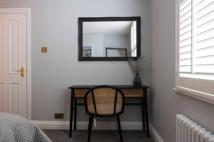 A stylish black-framed mirror hangs above a black desk with a rattan chair, positioned next to a white door and a window with white plantation shutters, all against light grey walls.