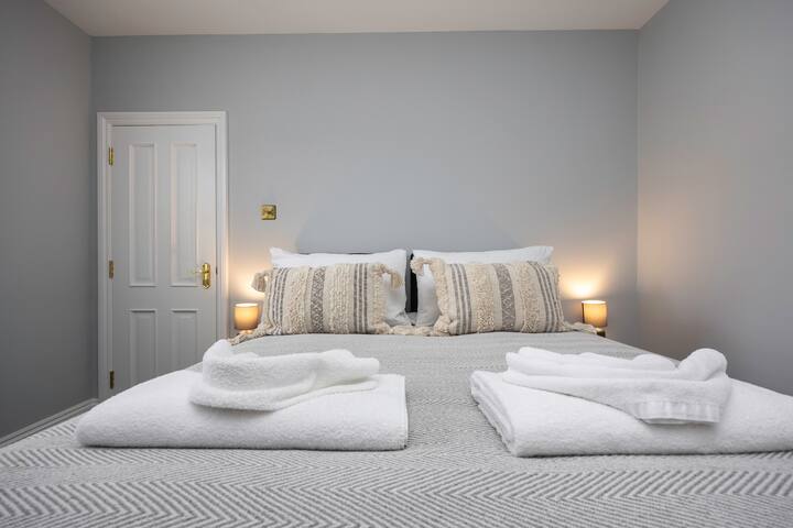A bright bedroom featuring a king-sized bed with a grey chevron-patterned duvet, white towels neatly folded, and two patterned throw pillows, flanked by bedside lamps against light grey walls.