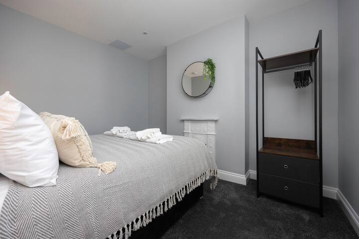 A compact bedroom with a single bed covered in a grey chevron blanket, a simple round mirror above a decorative white fireplace, and an open metal wardrobe on dark grey carpet.