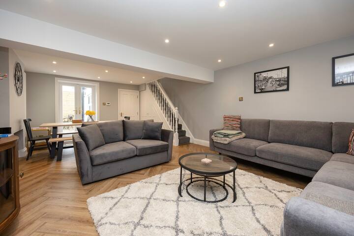 An open-plan living and dining area featuring a large gray sectional sofa and a smaller gray sofa on a white shag rug, facing a round coffee table, with a dining table and bench visible in the background, and stairs leading upstairs.