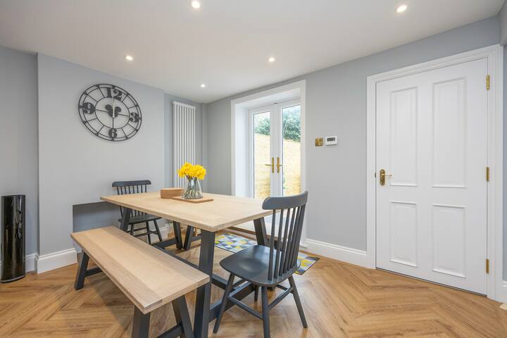 A dining area with light gray walls and light wood herringbone-patterned flooring, featuring a light wooden dining table with a bench and two dark gray chairs. A large black metal clock adorns the wall, and French doors lead to an outdoor area.