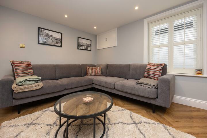 A corner of the living room showcasing a large gray sectional sofa adorned with striped throw pillows, a round glass coffee table, and a white shag rug, set on herringbone wood floors with light gray walls and a window with white blinds.