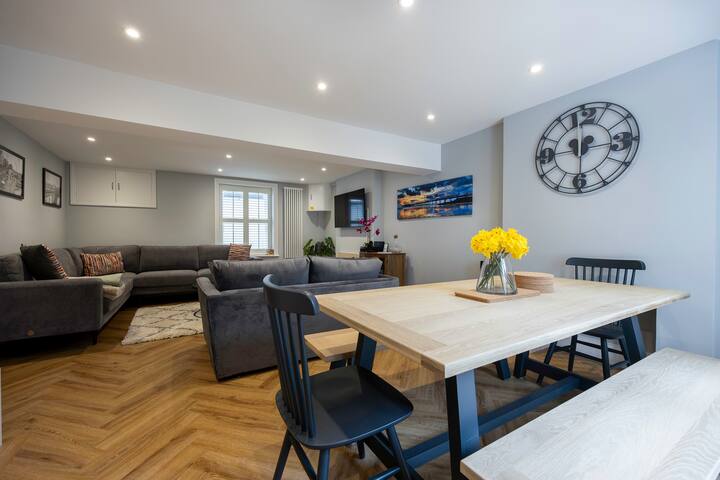 A spacious open-concept room featuring a dining area with a light wood table and black chairs/bench, flowing into a living area with gray sofas and a coffee table, all on herringbone wood floors, with a large wall clock.